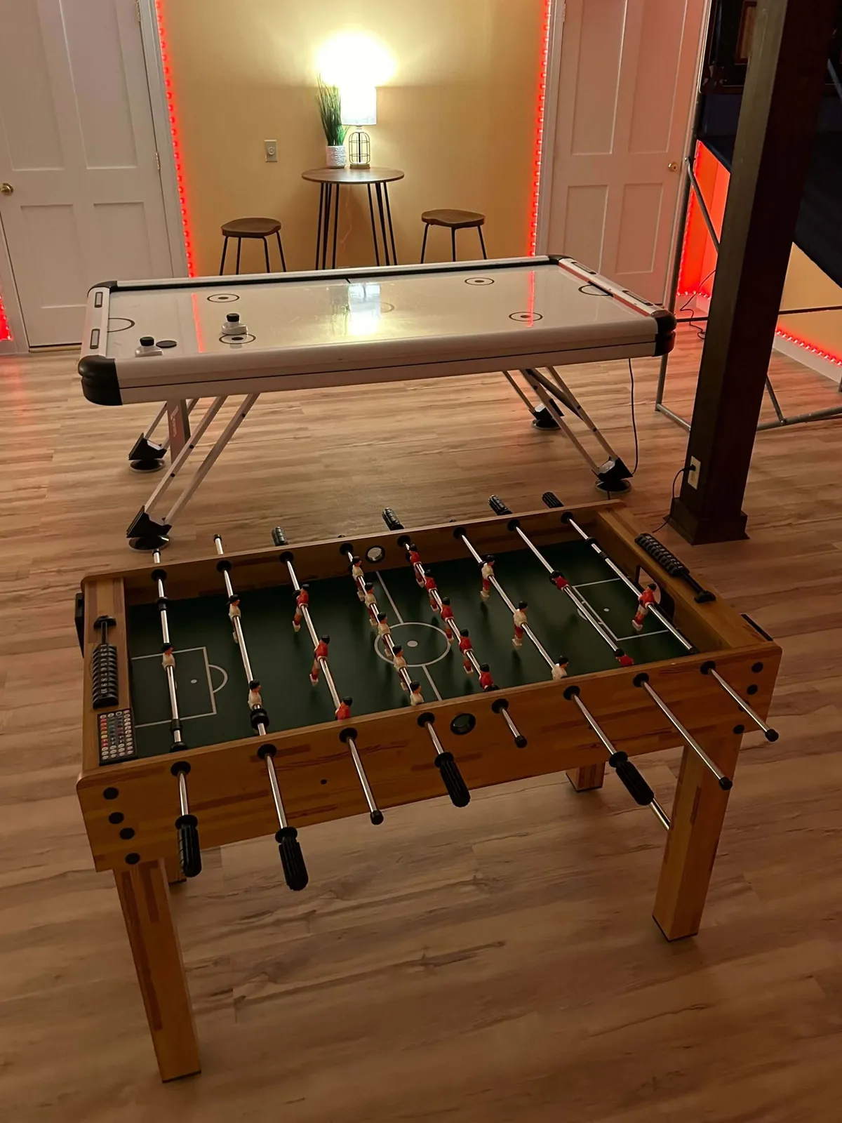 A vibrant game room with a foosball table, air hockey table, bar stools, and modern lighting, designed for friendly competition and socializing. The setup is casual and lively, echoing the fun atmosphere described for retreat comforts at home.