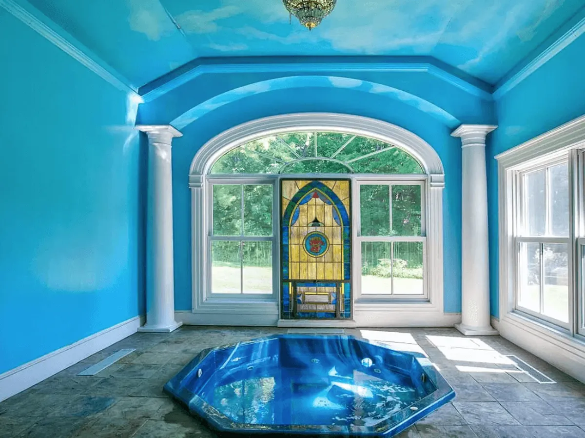 A serene octagonal hot tub set against blue walls, illuminated by natural light flowing through a stained glass window. The setting is peaceful and designed for relaxation and rejuvenation, emphasizing the luxurious spa-like experience.