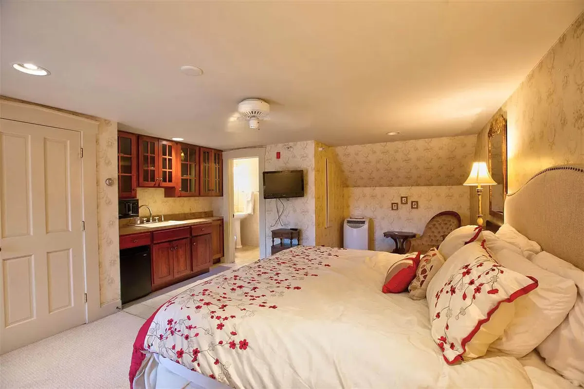 A warm and intimate suite featuring a king-sized bed with decorative pillows, a cozy kitchenette area, a wall-mounted TV, and a bedside lamp. The setting feels both luxurious and comfortable, with soft lighting and inviting decor that speaks to restful evenings and private relaxation.