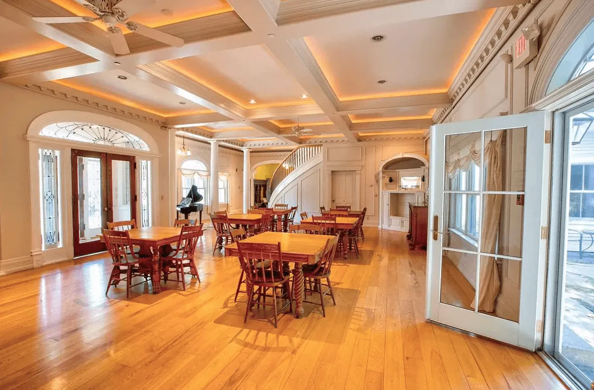 A bright dining area featuring wooden tables, large windows letting in sunlight, and a visible staircase. The communal setup encourages group dining and celebrations, capturing the essence of togetherness for milestones, shared meals, and festive occasions.