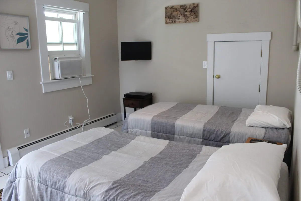 A relaxed bedroom with two beds, a wall-mounted TV, framed artwork, and an air conditioning unit. The layout supports shared occupancy while still offering individual comfort and entertainment.