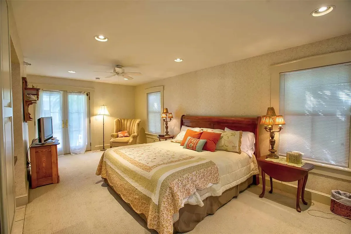 A cozy bedroom with a decorative quilt on the bed, an armchair, ambient lamps, and a television. Sunlight streams softly through the window, creating a peaceful atmosphere ideal for leisurely mornings or relaxing evenings.
