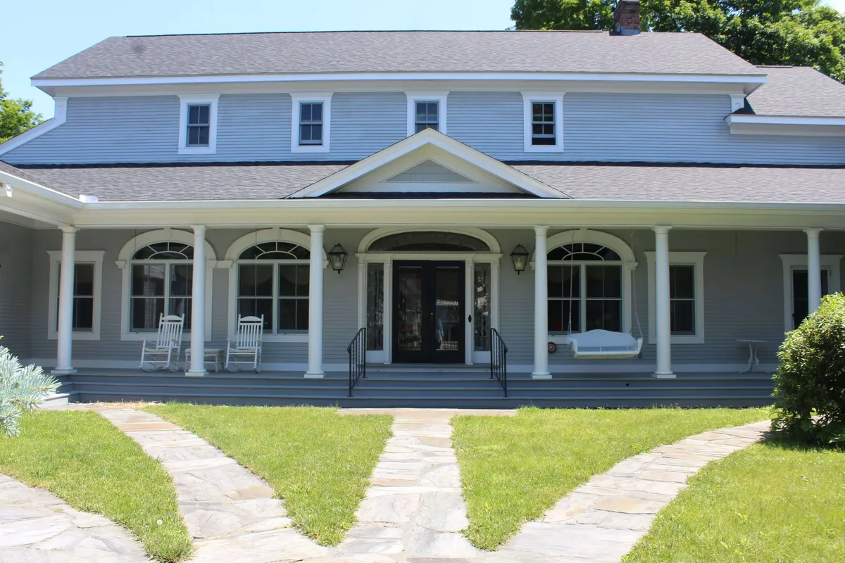 A wide exterior shot of the front porch with rocking chairs, arched windows, a stone pathway, and a well-tended lawn. The mood is cozy and inviting, ideal for family gatherings or relaxing after a day of local adventures.