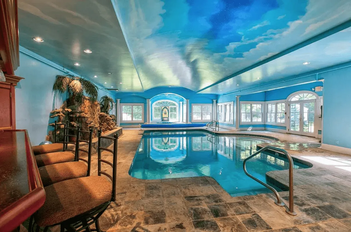 The image showcases a stunning indoor pool area with a sky-painted ceiling that evokes the feeling of a serene oasis. There's a chic seating bar along the pool, inviting guests to enjoy drinks and conversation, while an elegant water feature adds to the tranquil ambiance. The space is designed for both relaxation and entertaining, perfect for unwinding after a day in Vermont or hosting memorable gatherings.