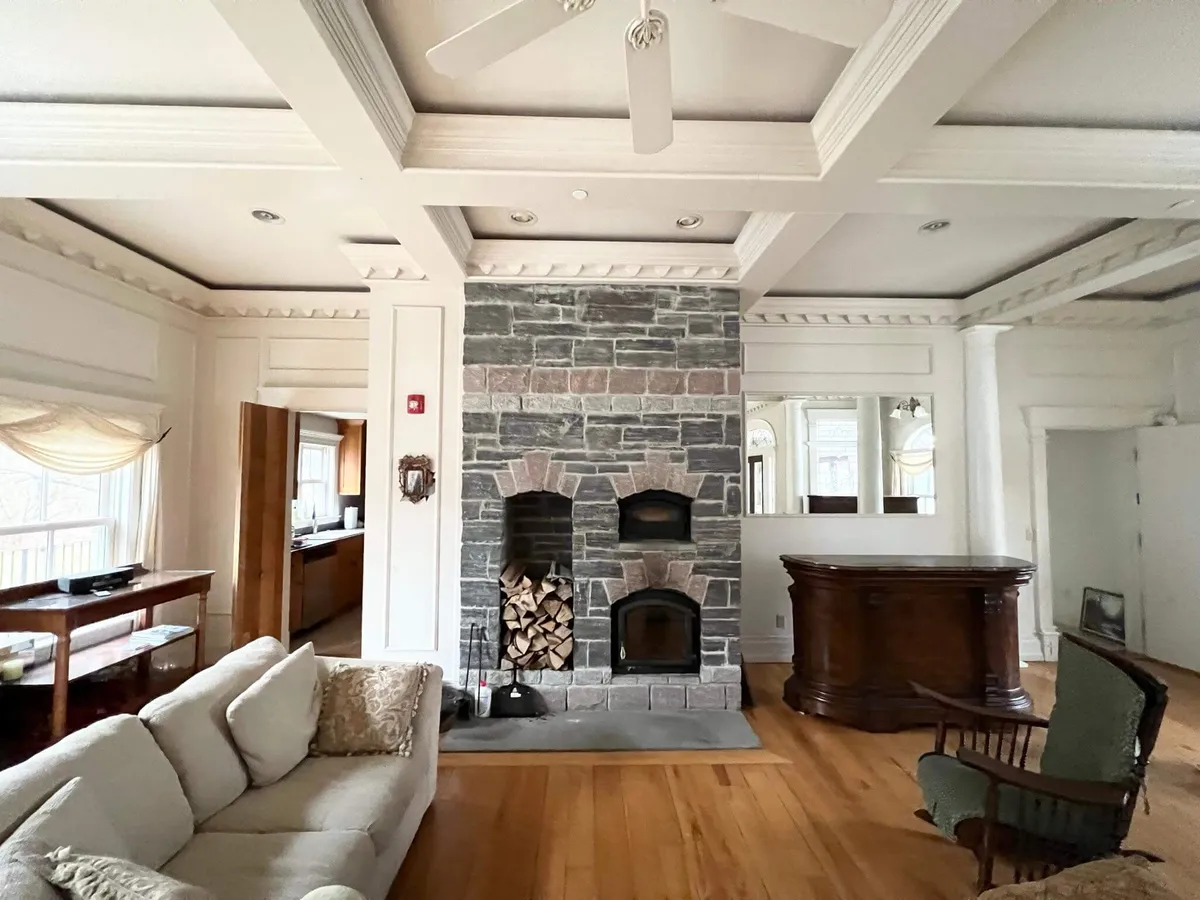 A cozy living area with a stone fireplace, sofa, wooden bar, and curtained window. The space is arranged for both intimate family gatherings and entertaining, with plush lounge seating and a warm, welcoming mood.