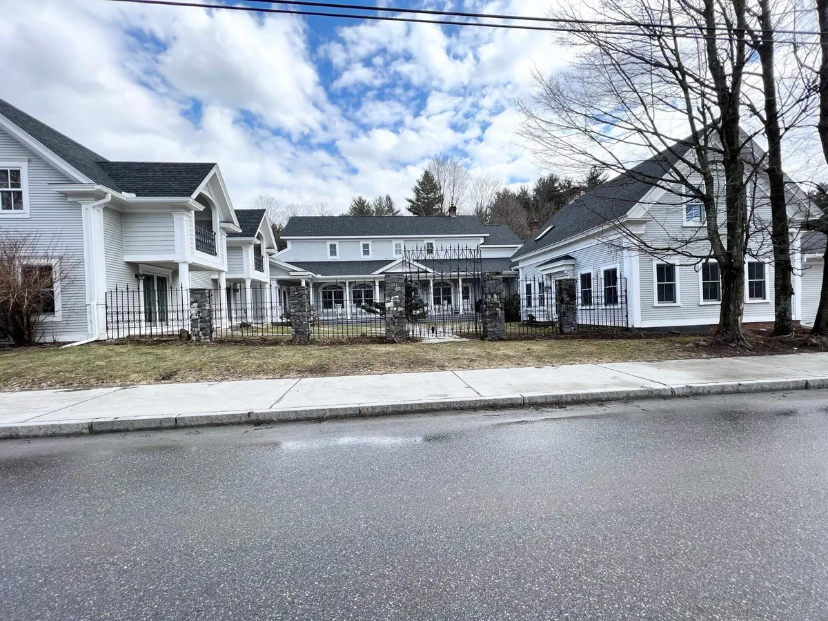 This image shows the grand main entrance of the property, with detailed architecture, a surrounding fence, lush landscaping, and a tree-lined street. The scene exudes a sense of arrival, community, and connection to Vermont’s natural beauty.