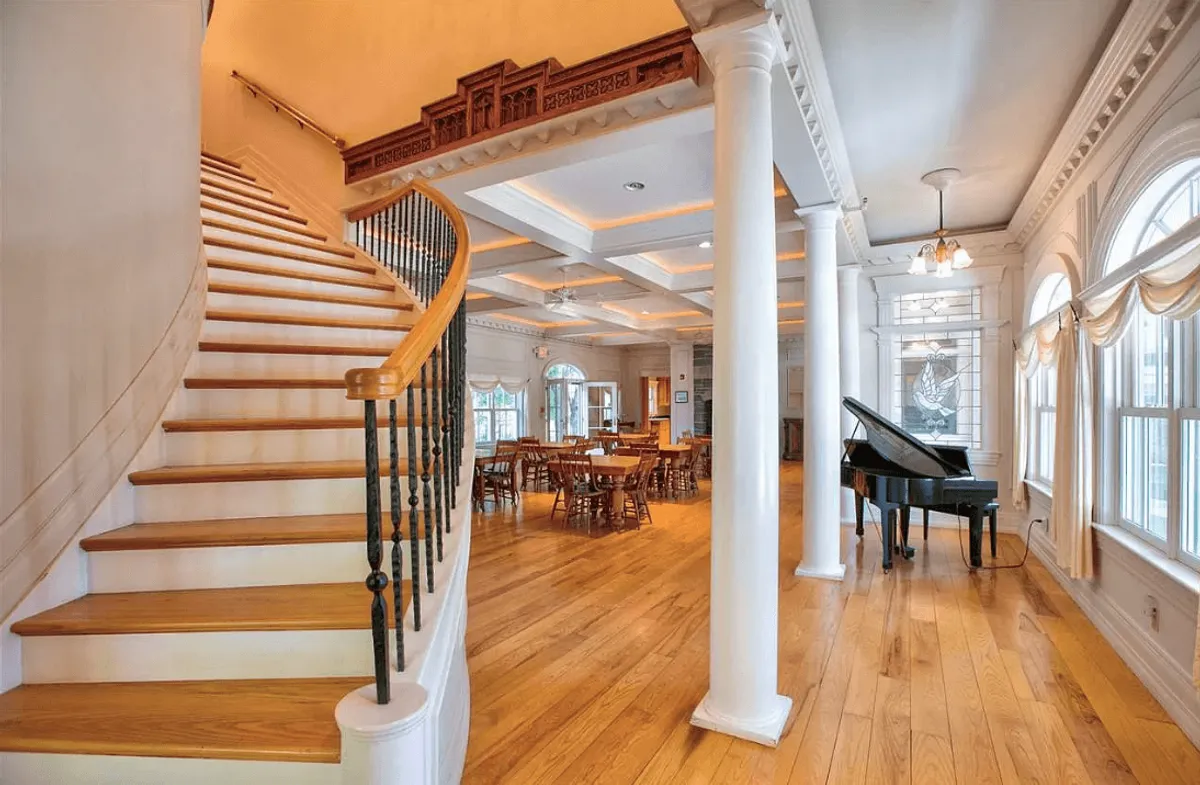 This image showcases a spacious, elegant interior with a staircase, grand piano, dining area, and large windows. The layout encourages social gatherings, with spaces for musical performances, festive meals, and cozy conversations. The abundance of natural light and sophisticated furnishings highlight a welcoming and upscale atmosphere.